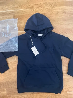 Mörkblå hoodie - Snygg mörkblå hoodie med broderad  på bröstet. Klassisk känguruficka framtill, justerbar dragsko i huvan och ribbade muddar. Tillverkad i mjuk bomull för en skön och avslappnad look. Perfekt för dig som vill ha en stilren och exklusiv hoodie.