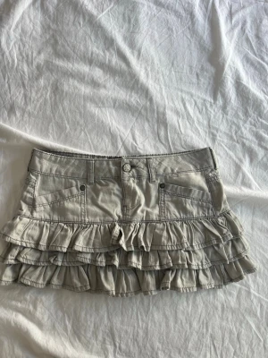 Beige volangkjol med fickor - Snygg beige kortkjol med flera volanglager och praktiska fickor fram. Kjolen har bälteshällor och en dekorativ patch bak. Perfekt för dig som gillar en lekfull och trendig stil.