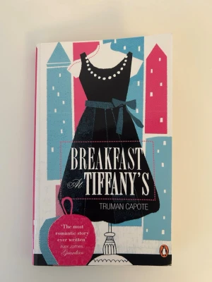 Breakfast at Tiffany's av Truman Capote - En ikonisk roman av Truman Capote, utgiven av Penguin. Omslaget är färgstarkt med svart, blått och rosa, och pryds av en elegant klänning mot en stadssilhuett. ISBN 978-0-241-95145-3. Boken är på engelska och är i mycket bra skick. 