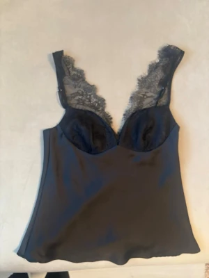Viral Zara topp  - Snygg svart topp  med djup urringning och spetsdetaljer vid axelbanden. Tillverkad i ett glansigt satinliknande material som ger en lyxig känsla. Perfekt för dig som vill ha något extra under kläderna. Viral Zara topp som är helt slutsåld passar storlek S/M
