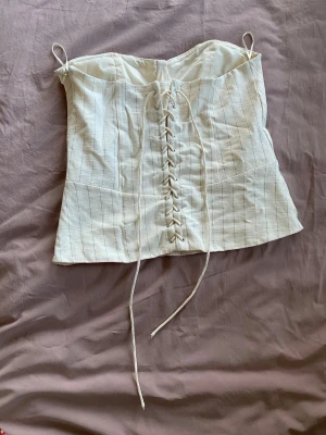 Vit korsett med snörning fram - Säljer en snygg vit korsett med tunna vertikala ränder och snörning framtill. Modellen har formade kupor och smala axelband för en feminin siluett. Materialet känns som bomullsmix och ger en klassisk look. Perfekt att styla till jeans eller kjol.