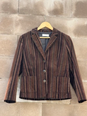 Randig brun kavaj i manchester C&A - Snygg kavaj i brun manchester med tunna vertikala ränder i beige, svart och vinrött. Klassisk krage, tre knappar framtill och två stora fickor. Fodrad insida och rak passform ger en cool retrovibe. 