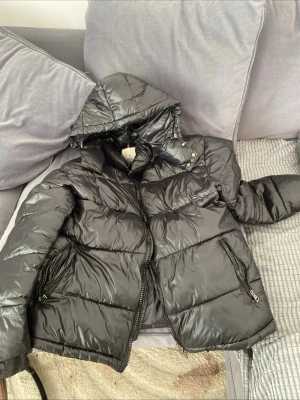 Svart pufferjacka från Moncler - Snygg svart pufferjacka från Moncler med glansig finish och avtagbar huva. Jackan har två dragkedjefickor framtill och quiltad design som håller värmen. Perfekt för kalla dagar och har klassisk Moncler-logga på insidan.