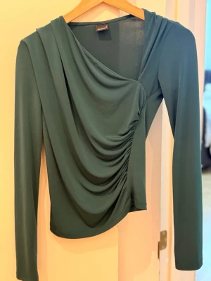 Mörkgrön draperad topp med lång ärm - Snygg mörkgrön topp med draperad front och lång ärm. Toppen har en asymmetrisk omlott-look och är tillverkad i ett mjukt, stretchigt syntetmaterial. Perfekt för dig som gillar trendiga detaljer och en unik siluett.
