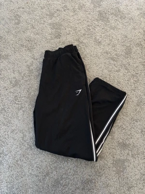 Svarta träningsbyxor från Gymshark - Säljer ett par svarta träningsbyxor från Gymshark med vita ränder längs benen och elastisk midja. Byxorna är tillverkade i ett ventilerande meshmaterial och har en diskret Gymshark-logga på låret. Perfekta för gymmet eller löprundan. Använda 1 gång köpta för 599kr