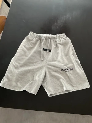 Grå Essentials Fear of God shorts - Snygga ljusgrå shorts från Essentials Fear of God med svart logga på benet. De har elastisk midja med snörning och är tillverkade i mjuk bomull, perfekta för en chill och avslappnad stil.
