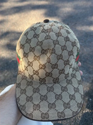 Gucci keps med GG-mönster - Snygg beige keps från Gucci med klassiskt brunt GG-monogram över hela kepsen. På sidan finns den ikoniska gröna och röda randen. Kepsen har böjd skärm och är tillverkad i canvasmaterial.