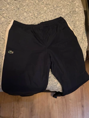 Marinblå shorts från Lacoste - Snygga marinblå shorts från Lacoste med vita sidopaneler och klassiska krokodilloggan på vänster ben. Shortsen har elastisk midja och är perfekta för sportiga aktiviteter eller en avslappnad stil. Tillverkade i ett lätt syntetmaterial.