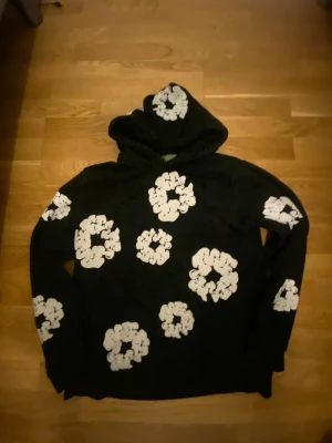 Denim Tears hoodie  - Säljer en svart hoodie med stora vita blommönster över hela tröjan, även på huvan. Hoodien har en avslappnad passform och är tillverkad i mjukt material. Perfekt för dig som gillar streetwear och vill sticka ut lite extra.