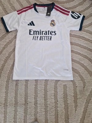 Real Madrid matchtröja Adidas vit - Snygg vit Real Madrid matchtröja från Adidas med klubbmärke och sponsortryck på bröstet. Tröjan har mörkblå detaljer vid ärmslut och hals, samt vinröda ränder på axlarna. Tillverkad i lätt och ventilerande material, perfekt för fotbollsträning eller match.