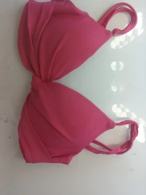 Rosa bikiniöverdel med twistad detalj - Snygg rosa bikiniöverdel med twistad front och dubbla axelband. Bikinin har vadderade kupor för extra komfort och stöd. Perfekt för strandhäng eller poolparty. Materialet känns mjukt och stretchigt för en skön passform.
