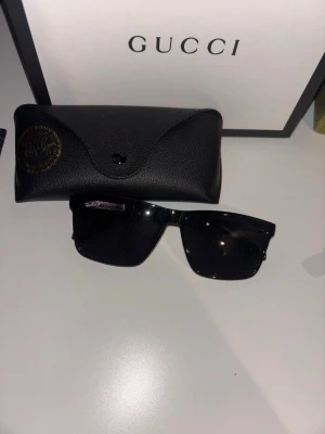 Svarta solglasögon från rayban  - Snygga svarta solglasögon från rayban med rektangulära bågar och mörka glas. Kommer med ett exklusivt svart fodral i skinn. Perfekta för dig som vill ha en stilren och lyxig accessoar till din outfit.