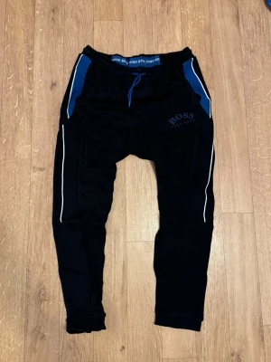 Svarta mjukisbyxor från Hugo Boss - Snygga svarta mjukisbyxor från Hugo Boss med blå detaljer och vita ränder längs benen. Byxorna har dragsko i midjan och fickor på sidorna. Perfekta för chill dagar eller när du vill ha en sportig look.