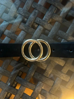 Svart bälte med dubbelring-spänne - Snyggt svart bälte med ett stilrent dubbelring-spänne i guld. Bältet är smalt och passar perfekt till jeans eller kjol. Klassisk design som ger en trendig touch till din outfit.