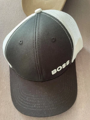 Svart och vit keps från BOSS - Snygg svart och vit keps från BOSS med broderad logga på framsidan. Kepsen har böjd skärm och vit mesh baktill för extra ventilation. Justerbar passform med klassisk trucker-modell. Perfekt för en avslappnad och trendig stil.