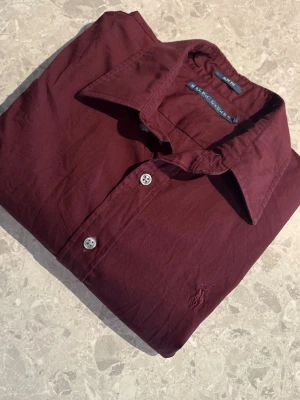 Vinröd skjorta  Ralph Lauren -  Kan prutas. Snygg vinröd skjorta från Ralph Lauren Skjortan  är i slim fit-modell. Perfekt för dig som gillar stilrena skjortor och inte vill sticka ut för mycket. Säljer skjortan föe att den inte används, använd några enstaka gånger. 
