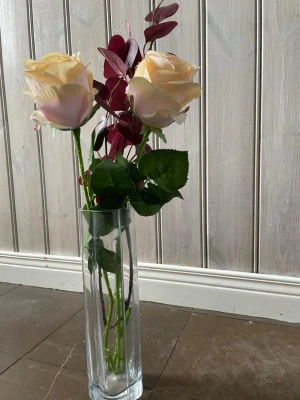 Dekorativ vas med konstblommor - Säljer en hög, rektangulär glasvas med konstgjorda blommor. Buketten består av två stora ljusgula rosor, mörkröda blad och gröna blad. Perfekt som dekoration i hemmet för en stilren och elegant känsla.  Ca 29 cm lång 