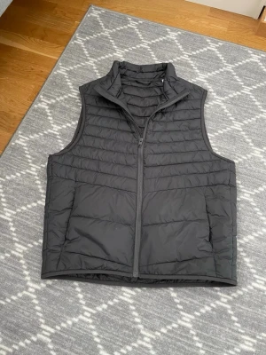 Mörkgrå Jack n Jones vest - Säljer en mörkgrå dunväst med quiltad design och hög krage. Västen har dragkedja framtill och två stora fickor. Perfekt för lager-på-lager och håller dig varm under kyliga dagar. Materialet är lätt och smidigt, vilket gör den enkel att bära.