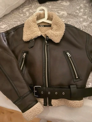 Brun skinnjacka med fuskpäls från Zara - Snygg brun skinnjacka från Zara med krage och detaljer i beige fuskpäls. Jackan har bälte i midjan, dragkedjor på fickorna och ärmarna samt cool bikerstil. Perfekt för dig som vill ha en trendig och varm look. Passar xs/s
