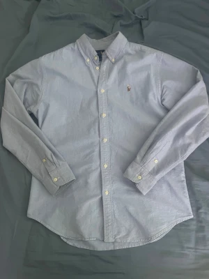 Ljusblå skjorta från Ralph Lauren - Klassisk ljusblå skjorta från Ralph Lauren med button-down krage och broderad logga på bröstet. Skjortan har långa ärmar, vita knappar och är tillverkad i mjuk bomull. Perfekt nu till sommaren!