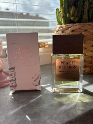 Peach Macaron parfym - Peach Macaron från Theodoros Kalotinis är en elegant parfym i en genomskinlig, fyrkantig glasflaska med träfärgat lock och guldbrun etikett. Doften kommer i en vit kartong med guldtext och illustration av macarons. Perfekt för dig som gillar fruktiga och söta dofter. 50ml . Ny 