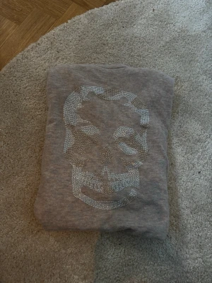 Grå stickad tröja med döskalle - Säljer en beige stickad tröja med en döskalle framtill i silvriga stenar/nitar. Tröjan har lång ärm och rund halsringning. Tröjan är köpt i new york, och är ifrån zadig & voltaire.
