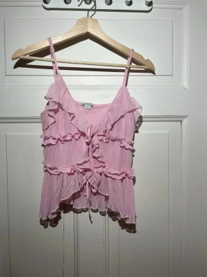 Rosa volangtop från Amisu - Söt rosa linnetopp från Amisu med tunna axelband, volangdetaljer och knyt framtill. Toppen har en luftig och lätt struktur, perfekt för varma dagar. Materialet känns mjukt och toppen har en feminin look med flera lager volanger.