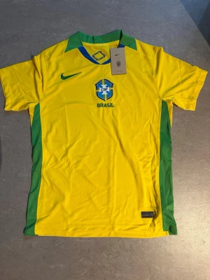 Brasilien fotbollströja Nike gul/grön - Snygg Brasilien fotbollströja från Nike i gult med gröna detaljer och klassisk CBF-logga på bröstet. Tröjan har korta ärmar, grönt krageparti och är tillverkad i lätt, ventilerande material som passar perfekt till träning eller match.