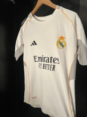 Real Madrid matchtröja Adidas vit - Säljer en vit Real Madrid matchtröja från Adidas med klubbmärke och sponsortryck på bröstet. Tröjan har svarta detaljer, gula sömmar och Champions League-märke på ärmen. Tillverkad i lätt och ventilerande material, perfekt för fotboll eller träning.