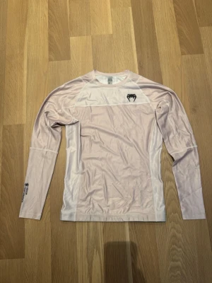 Venum Biege långärmad  - Säljer en Beige långärmad rashguard från Venum med svart logga på bröstet och text på ryggen. Tröjan är tillverkad i ett stretchigt, funktionsmaterial som andas och har vita paneler på sidorna. Perfekt för träning eller kampsport.