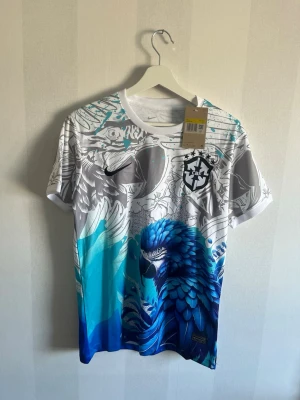 Nike Brasil T-shirt med blå papegoja - Unik Nike t-shirt för Brasilien med ett stort blått papegojmotiv och grafiska detaljer i vitt, grått och blått. Kortärmad modell med rund hals och tryckt CBF-logga på bröstet. Tillverkad i lätt och skönt material, perfekt för dig som vill sticka ut.