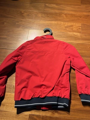 Röd vindjacka från North82 - Snygg röd vindjacka från 8848 med svartvita muddar och dragkedja framtill. Jackan har tryck på bröstet med texten 'Blue Water Bridge 1982' och två sidofickor. Materialet är tunt och vindtätt, perfekt för blåsiga dagar.