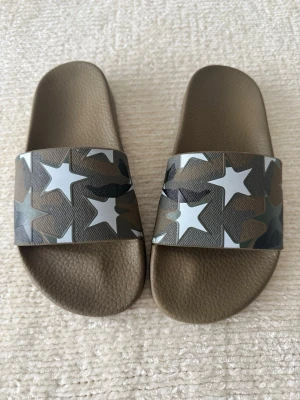 Olivgröna tofflor med stjärnmönster - Säljer ett par olivgröna tofflor med bred rem i camomönster och vita stjärnor. Tofflorna har en klassisk slip-in design och är tillverkade i syntetmaterial med struktur på fotbädden för extra komfort. Perfekta för chill dagar hemma eller på stranden.