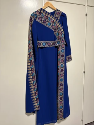 En palestinsk klänning  - Unik blå långklänning med färgglada broderade mönster längs ärmar, halsringning och framstycke. Klänningen har lång ärm och ett traditionellt, elegant snitt. Perfekt för dig som vill sticka ut med vackra detaljer och färg.