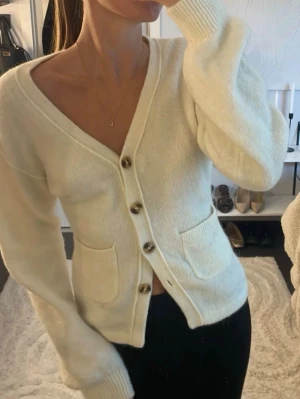 Vit kofta med knappar - Mysig vit kofta med v-ringning och fyra bruna knappar framtill. Koftan har två praktiska fickor fram och ribbade muddar vid ärmslut och nederkant. Tillverkad i ett mjukt, stickat material som känns skönt mot huden.Liten fläck på armen som nog går bort i tvätten, annat superbra skick.