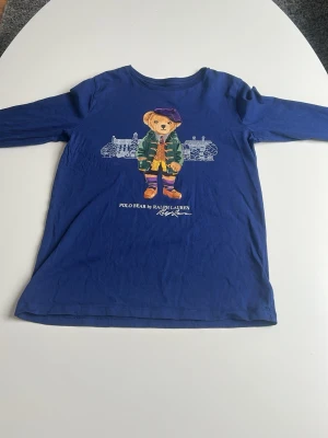 Blå långärmad t-shirt Polo Ralph Lauren - Säljer en blå långärmad t-shirt från Polo Ralph Lauren med ikoniska Polo Bear-trycket framtill. T-shirten är i mjuk bomull och har en klassisk passform. Perfekt för dig som gillar stilrena plagg med en twist!
