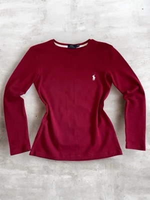 Vinröd långärmad topp från Polo Ralph Lauren - Klassisk vinröd långärmad topp från Polo Ralph Lauren med rund halsringning och ikonisk vit broderad logga på bröstet. Tillverkad i mjuk bomull som känns skön mot huden. Perfekt att styla med jeans eller kjol för en clean look.