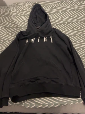 Svart hoodie från Amiri - Säljer en svart hoodie från Amiri med stor vit logga över bröstet. Hoodien har en klassisk känguruficka framtill och en rymlig huva. Perfekt för dig som gillar streetwear och vill ha något stilrent men ändå med attityd.