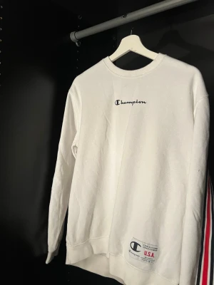 Vit stickad sweatshirt från Champion - Säljer en vit stickad sweatshirt från Champion med broderad logga på bröstet och en patch vid nederkanten. Tröjan har rund halsringning och långa ärmar. Perfekt för en avslappnad och sportig stil. 