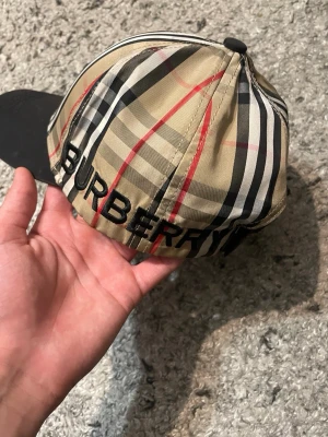 Burberry keps med klassiskt rutmönster - Snygg keps från Burberry med det ikoniska beige, svarta och röda rutmönstret. Framsidan har en svart patch med texten 'BURBERRY LONDON ENGLAND' och baksidan har broderad logga. Skärmen är svart och materialet känns slätt och tåligt.