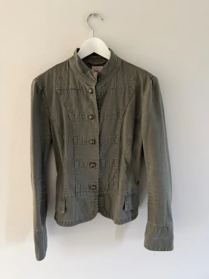 Militärinspirerad jacka Colours of the World - Snygg militärgrön jacka från Colours of the World med ståkrage och dekorativa knappar framtill. Storlek 38 som passar xs-m och är i bra skick.