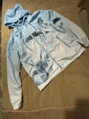 Ljusblå vindjacka med örntryck - Ljusblå vindjacka med huva och dragkedja framtill. Jackan har stora tryck av örnar i flykt både fram och bak samt texten på ryggen. Två fickor med dragkedja och justerbar nederkant. Materialet är tunt och glansigt, perfekt för blåsiga dagar.