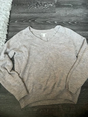 Ljusgrå/beige v-ringad stickad tröja - Mysig ljusgrå stickad tröja med v-ringning och långa ärmar. Tröjan har ribbade muddar vid ärmslut och nederkant, vilket ger en snygg och avslappnad look. Perfekt att matcha med jeans eller kjol. Lite nopprig.