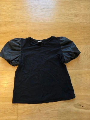 Svart topp med puffärmar - Trendig svart topp med puffiga korta ärmar i glansigt material. Klassisk rund halsringning och rak passform. Perfekt för dig som vill ha en enkel men ändå statement topp till din outfit.