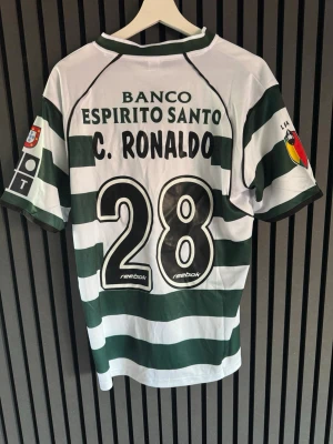 Sporting Lissabon matchtröja Reebok - Grönvit randig matchtröja från Sporting Lissabon med C. Ronaldo och nummer 28 på ryggen. Tröjan har klubbmärke på bröstet, sponsorloggor och korta ärmar. Tillverkad i lätt och ventilerande material, perfekt för fotbollsträning eller samling.