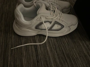 Vita Dior sneakers med grå detaljer - OBS KVITTO OCH KARTONG FINNS! Snygga vita Dior sneakers med grå och svarta detaljer. Skorna har en chunky sula och är tillverkade i mesh och läder för en modern look. Klassisk låg modell med snörning och diskret Dior-logga på sidan. Perfekta för dig som vill ha en clean och exklusiv stil. 