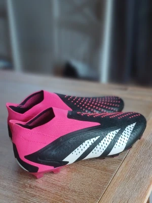 Adidas rosa och svarta fotbollsskor - Säljer ett par Adidas fotbollsskor i en grymt snygg färgkombination av rosa, svart och vitt. Skorna har en strumpliknande passform utan snörning, med gummidetaljer på ovandelen för bättre bollkontroll och klassiska Adidas-ränder på sidan. Perfekta för dig som vill sticka ut på planen. Storlek 42 2/3 