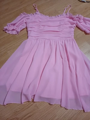 Ljusrosa kort klänning med volang - Supersöt ljusrosa klänning med volangdetaljer och rynkade partier upptill. Klänningen har korta puffärmar och tunna axelband, samt en luftig kjol i chiffongliknande material. Perfekt för sommarens alla tillfällen!