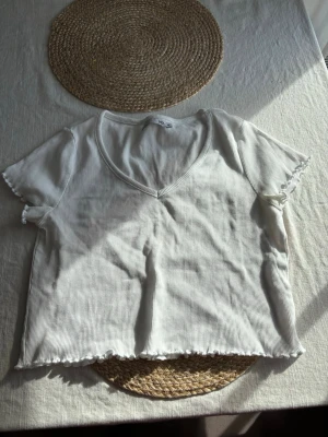 Vit ribbad croppad topp - Säljer en vit ribbad croppad topp med v-ringning och korta ärmar. Toppen har vågiga kanter vid ärmslut och nederkant för en söt touch. Perfekt till jeans eller kjol för en enkel men snygg look. Storlek M