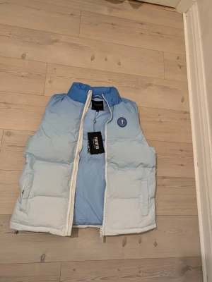 Ljusblå dunväst från Varsity - Snygg ljusblå dunväst från Varsity med vit dragkedja och hög krage. Västen har två fickor framtill och ett broderat Varsity-märke på bröstet samt stor text på ryggen. Perfekt för lager på lager och streetwear-stil.